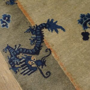Muted greenish-tan rug with blue stylized motifs and an orange accent stripe on wood flooring.