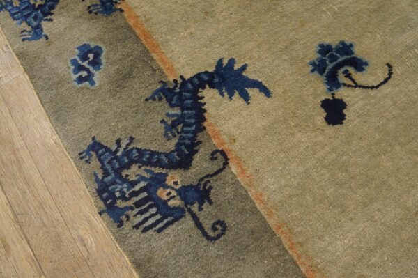 Muted greenish-tan rug with blue stylized motifs and an orange accent stripe on wood flooring.