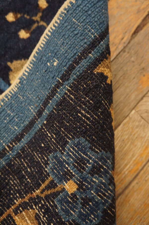 A navy and tan patterned rug is folded over, showing a blue and white woven back and resting on worn wood planks.