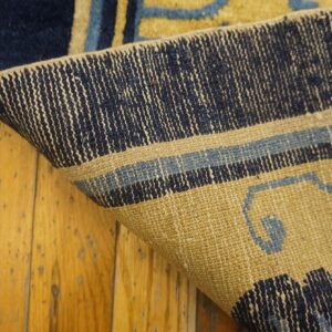 Curled corner of a blue and tan patterned rug reveals its backing structure over yellow wood planks.