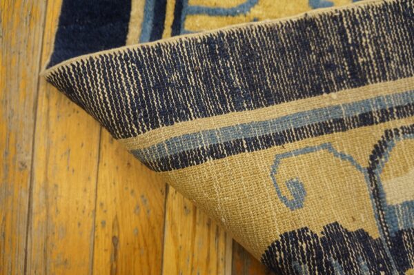 Curled corner of a blue and tan patterned rug reveals its backing structure over yellow wood planks.