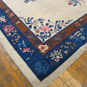 A light beige rug corner with blue and brown floral borders rests on a worn wood floor.