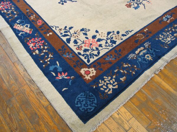 A light beige rug corner with blue and brown floral borders rests on a worn wood floor.