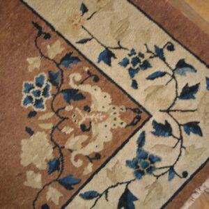 Corner detail of a low-pile rug showing brown field, cream border, and detailed blue floral pattern on wood planks.