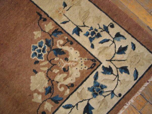 Corner detail of a low-pile rug showing brown field, cream border, and detailed blue floral pattern on wood planks.
