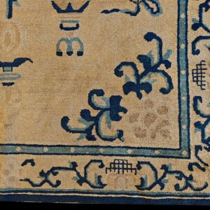 Corner of a tan rug with dark blue and light blue borders and stylized scrollwork designs.