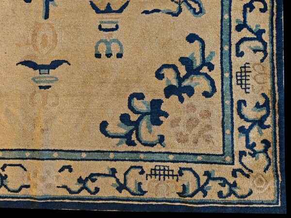 Corner of a tan rug with dark blue and light blue borders and stylized scrollwork designs.