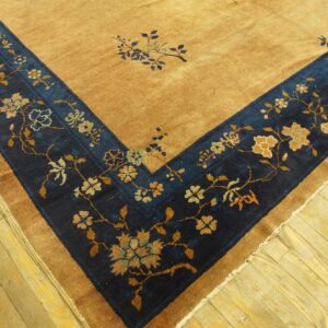 Corner of a tan rug featuring a wide, navy floral border against yellow hardwood flooring.