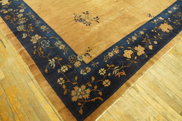 Corner of a tan rug featuring a wide, navy floral border against yellow hardwood flooring.