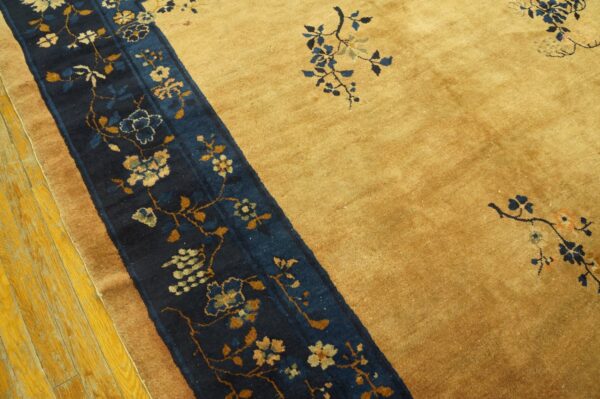 Gold rug with a wide navy blue border stripe featuring floral and vine motifs, sitting on yellow wood floor.
