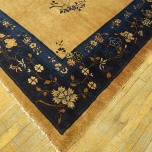 A golden-tan rug corner with a dark blue floral border rests on rustic yellow wood plank flooring.