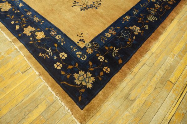 A golden-tan rug corner with a dark blue floral border rests on rustic yellow wood plank flooring.