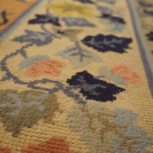 Close-up detail of a low-pile rug showing golden-tan ground with navy, gray, sage, and coral floral motifs.