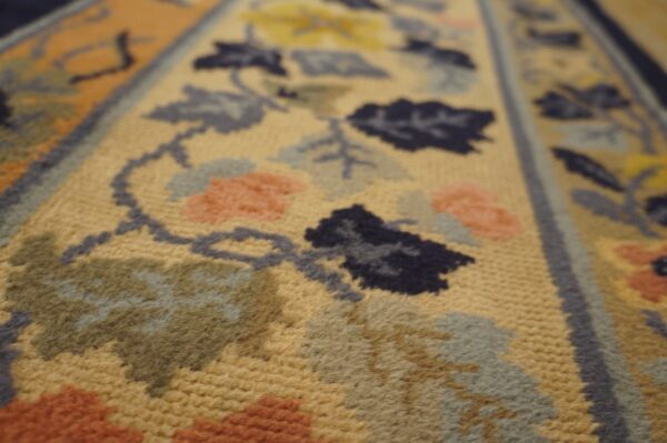 Close-up detail of a low-pile rug showing golden-tan ground with navy, gray, sage, and coral floral motifs.