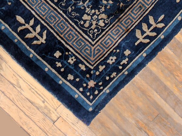 Navy blue rug with tan floral and geometric border patterns overlaps light wide-plank hardwood flooring.