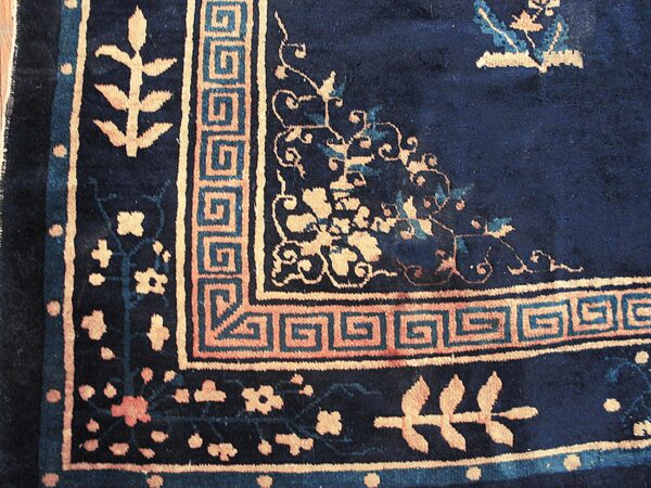 Navy blue rug corner featuring tan greek key and scrolling floral borders with slight teal accents.