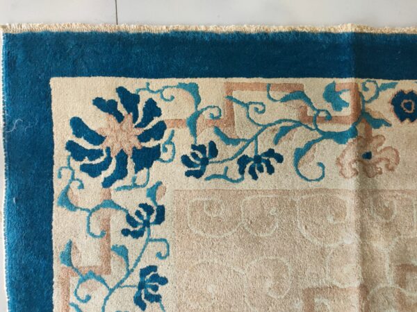 High-pile rug corner with a teal border, cream field, and pink and blue floral geometric pattern on white tile.