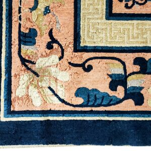 Thick pile rug detail showing peach field, navy border, and cream key-fret pattern in the corner.