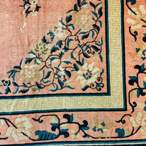 Corner detail of a pink rug featuring elaborate cream and teal scrolling floral motifs and patterned borders.