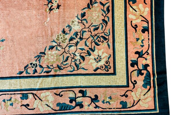 Corner detail of a pink rug featuring elaborate cream and teal scrolling floral motifs and patterned borders.
