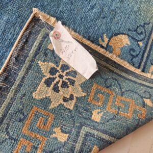 Close up of a blue and tan patterned rug, folded to show texture and an attached size tag.