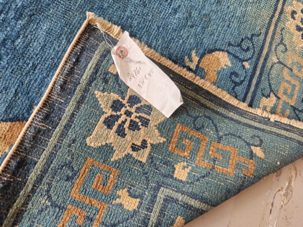 Close up of a blue and tan patterned rug, folded to show texture and an attached size tag.