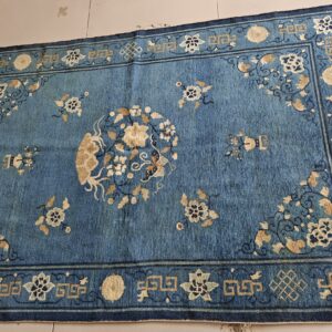 Distressed blue low-pile rug featuring a beige floral medallion and complex geometric border on light tiled flooring.