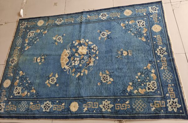 Distressed blue low-pile rug featuring a beige floral medallion and complex geometric border on light tiled flooring.