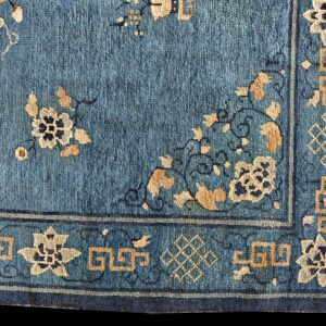 Blue, low-pile rug corner featuring tan floral designs and a border with geometric fret patterns.