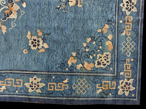 Blue, low-pile rug corner featuring tan floral designs and a border with geometric fret patterns.