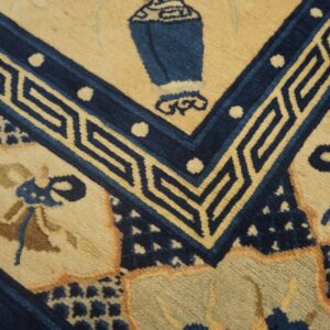 Close view of a tan and navy blue rug featuring a central urn motif and geometric key border.