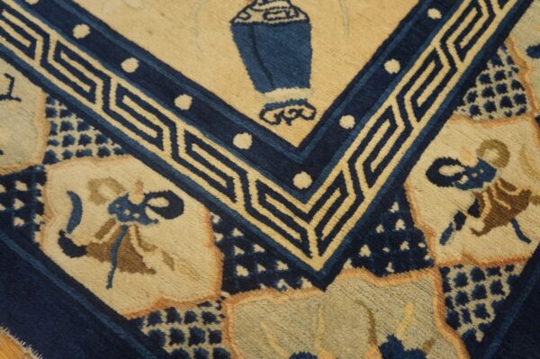 Close view of a tan and navy blue rug featuring a central urn motif and geometric key border.
