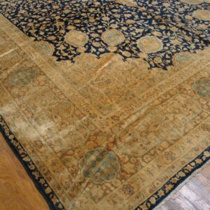 The corner of a dark blue and gold floral rug with a wide beige border lies on wood flooring.