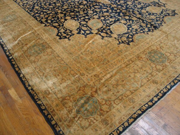 The corner of a dark blue and gold floral rug with a wide beige border lies on wood flooring.