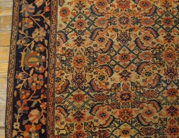 Close-up of a low-pile rug with dense orange, navy, and beige patterns, bordered by dark blue florals, on wood flooring.
