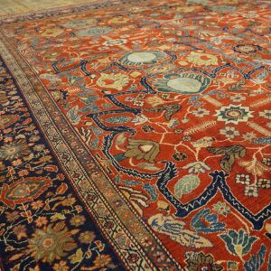 Ornate red-orange rug with a navy border and multicolor traditional floral patterns on hardwood flooring.