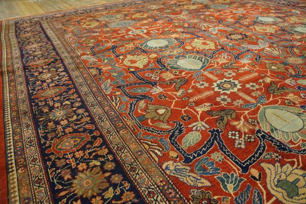 Ornate red-orange rug with a navy border and multicolor traditional floral patterns on hardwood flooring.