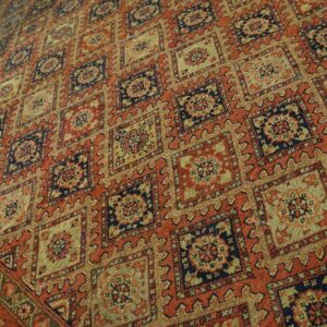 Close view of a woven rug with repeating geometric squares in red-orange, gold, and navy.