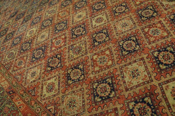 Close view of a woven rug with repeating geometric squares in red-orange, gold, and navy.