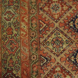 Detailed low-pile rug in rust, olive, and navy featuring repeating diamond patterns on light wood flooring.
