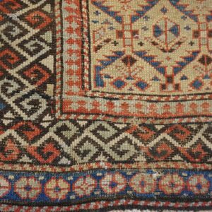 Geometric rug corner detail with tan field, diamond lattice, and floral borders in blue, red, and brown.
