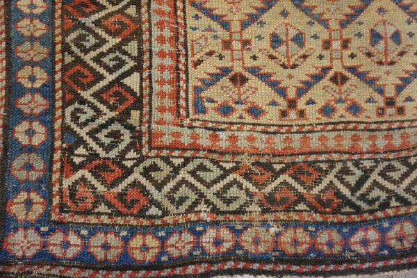 Geometric rug corner detail with tan field, diamond lattice, and floral borders in blue, red, and brown.
