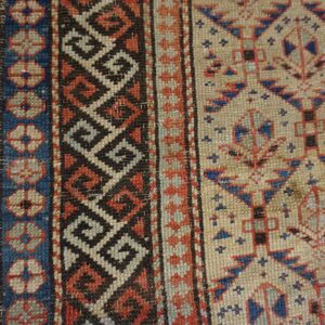Geometric rug detail with borders in blue, red, and black on a tan field, resting on light wood.