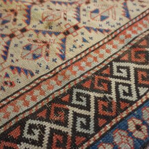 Low pile rug close-up showing beige field and bold borders with blue, red, and black geometric motifs.
