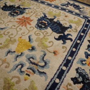 Textured pile rug with deep blue, orange, and green stylized patterns on a light field, bordered by navy trim.