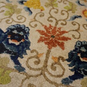 Textured rug detail with blue lion figures, rust flowers, and green scrolling vines on a cream background.