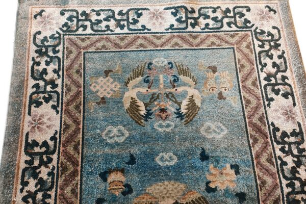 Close-up of a distressed blue rug featuring central cranes, floral patterns, and intricate multi-colored borders.