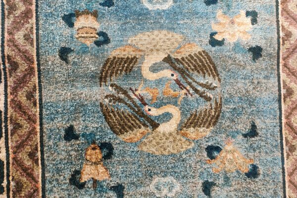 Low-pile, muted blue rug with a central design of two beige birds and a patterned border.