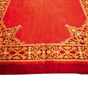 High-pile orange-red rug featuring elaborate gold foliate motifs along the patterned perimeter border.