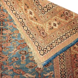 Close-up of a low-pile rug folded, showing the contrasting blue field and tan/coral patterned border.